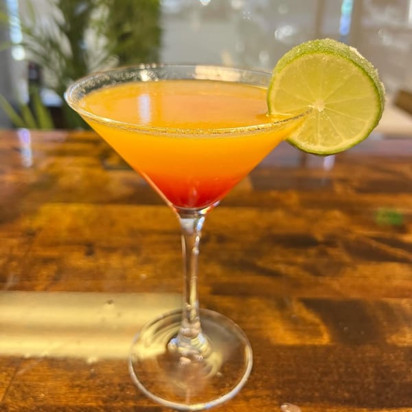 A vibrant orange and red cocktail in a martini glass, garnished with a sugar-rimmed lime slice.