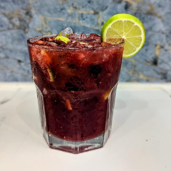 A Brazilian caipirinha cocktail, dark red with ice, lime, and fruit pieces, against a blue and white background.