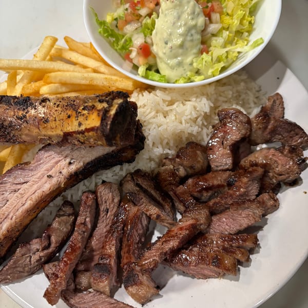Brazilian meal with grilled ribs, sliced steak, rice, fries, and a salad with dressing.